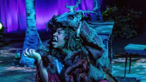 Actor wearing a cloak with an antlered skull crouches and looks stage right